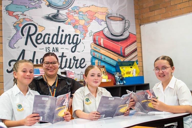 Photo shows three happy students reading in the library with their teacher.